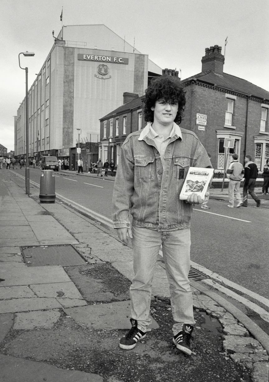 In 1991, photographer Richard Davis followed supporters from across the North West for a book about football, fashion and music. Richard spent much of the season taking pictures of fans from Liverpool, Everton and more for the book Football With Attitude. In collaboration with Lower Block, the Going to the Match zine has been published to coincide with an exhibition of the same name at Bury Art Museum, running from October 2024 to March 2025.
Pictured: Everton Fanzine seller Goodison Park, 1991