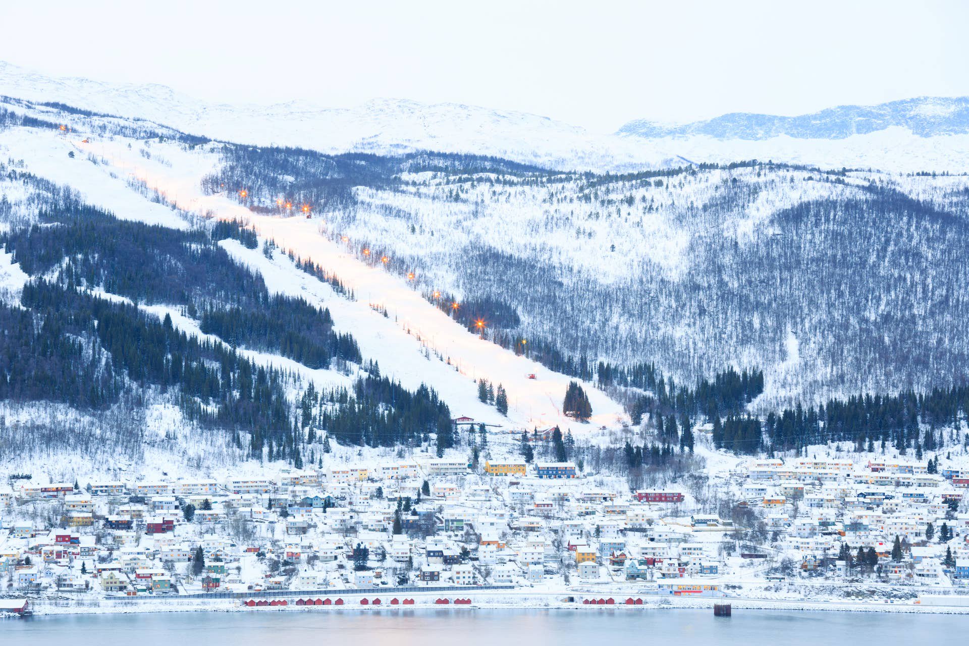 Verschneite Stadt Narvik mit beleuchteter Skipiste am Hang, umgeben von Bergen in Norwegen.
