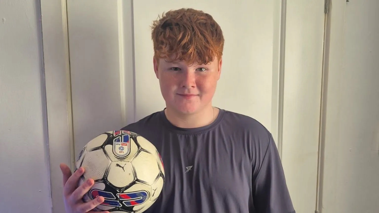 Handout Morgan-Lee stands in front of a white door and holds a football in one hand. He is wearing a grey t-shirt