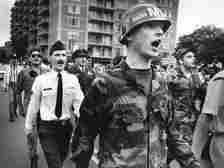 Men in military uniform from different eras walk down a street in Washington, D.C. in this black-and-white photo from the early 1990s. A multistory building is seen behind them.