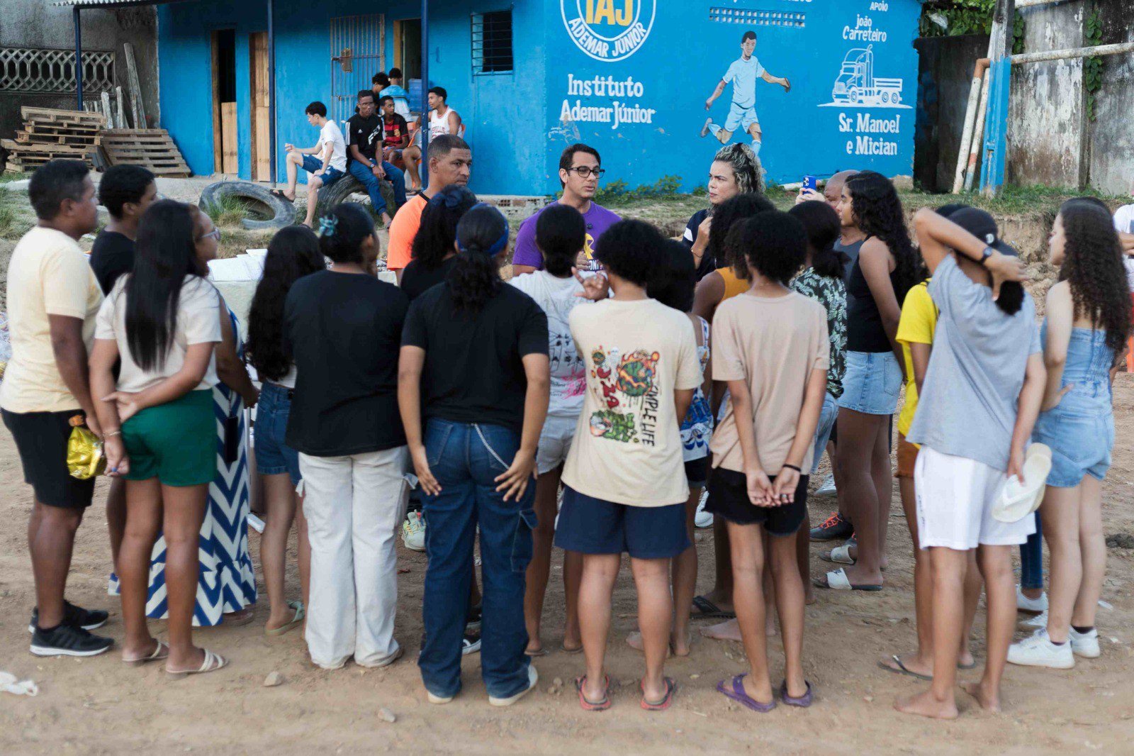 Um grupo de jovens e adultos, a maioria de costas, forma um círculo em frente a um prédio azul com a inscrição Instituto Ademar Junior e um desenho de jogador de futebol. Duas pessoas no centro do círculo, uma de óculos e camiseta roxa, e outra de cabelo loiro, parecem conversar com os demais. O chão é de terra batida, e ao fundo há mais pessoas sentadas e materiais de construção