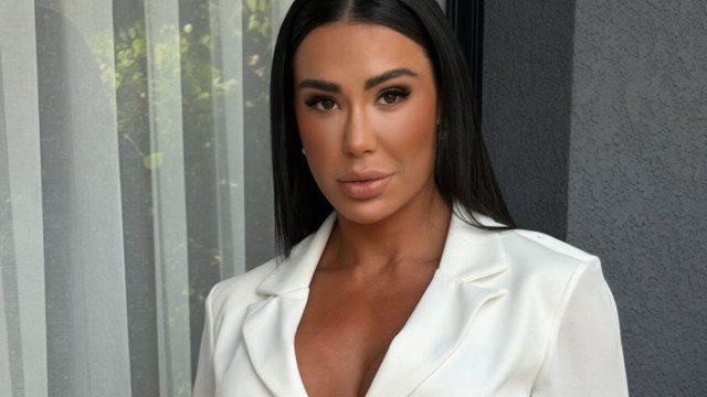 Mulher de pele bronzeada, cabelos pretos lisos e longos, maquiagem marcante, usando um blazer branco, olhando diretamente para a câmera com expressão séria. Ao fundo, cortinas brancas translúcidas e uma parede cinza texturizada.