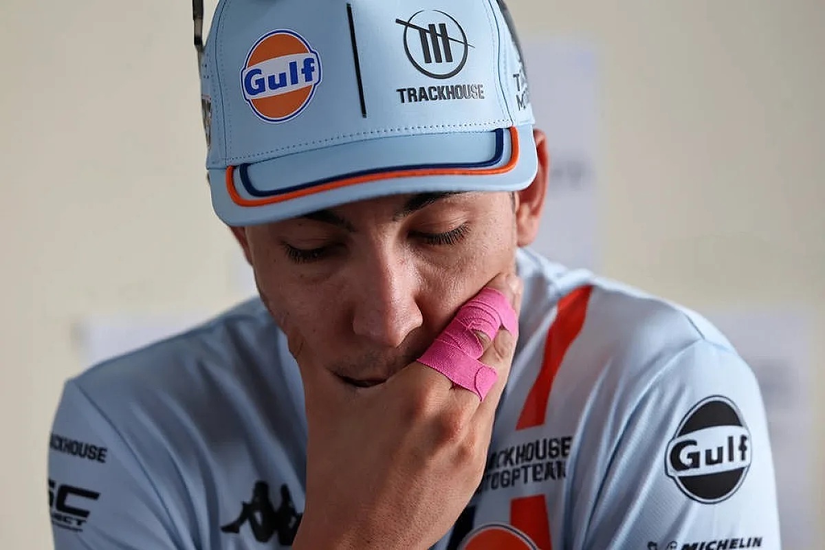 Rider in a light blue Gulf-branded cap and Trackhouse MotoGP shirt, with pink tape on his hand, looks down pensively in the paddock