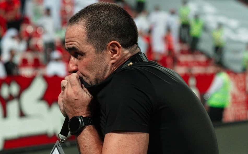 Homem de camiseta preta segura crachá pendente do pescoço, em estádio com público ao fundo.