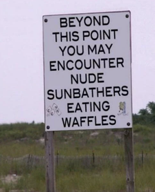 Sign warning: &quot;Beyond this point, you may encounter nude sunbathers eating waffles.&quot; The sign is placed on a beach