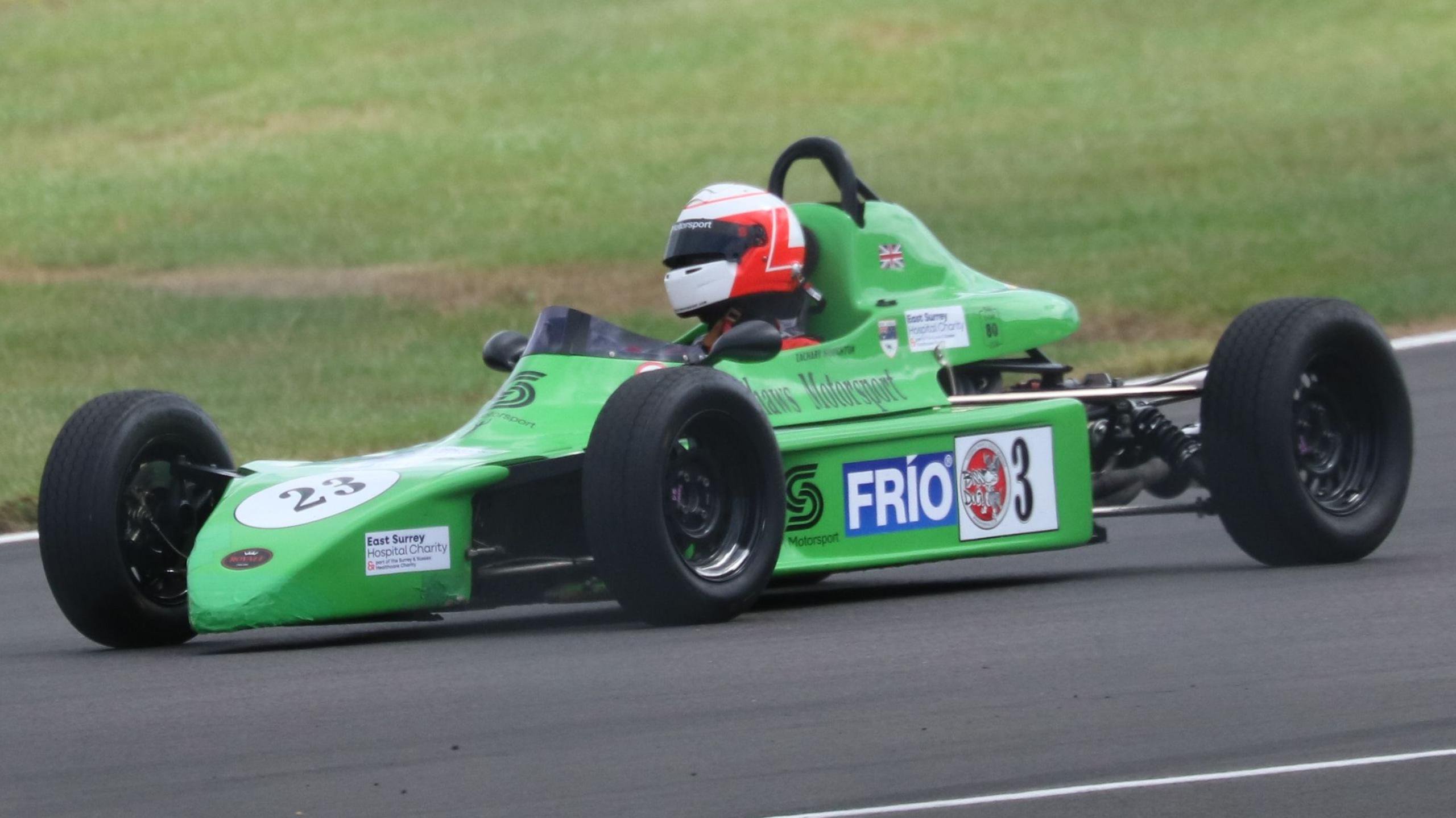 A green racing car on a track being driven by a person in an orange and white helmet.