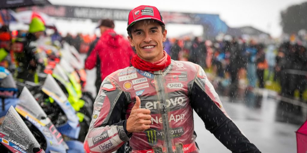 Rider in red Ducati leathers with a thumbs-up in the rainy MotoGP paddock, sponsor logos and a red cap visible