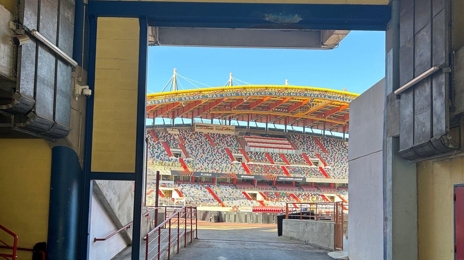 View from a stadium tunnel towards a football pitch and a modern stand with multi-coloured seating, red stairways and a curved yellow roof, under a clear blue sky.