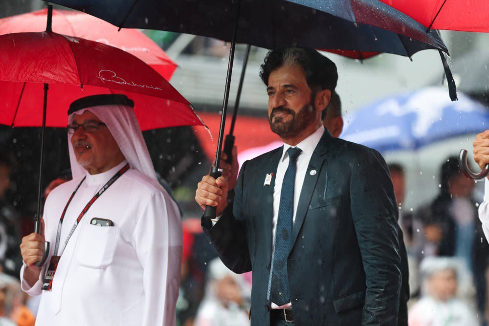 Two men under umbrellas in heavy rain at a motorsport event; the man in a dark suit is the article's subject, FIA president Ben Sulayem, per context.