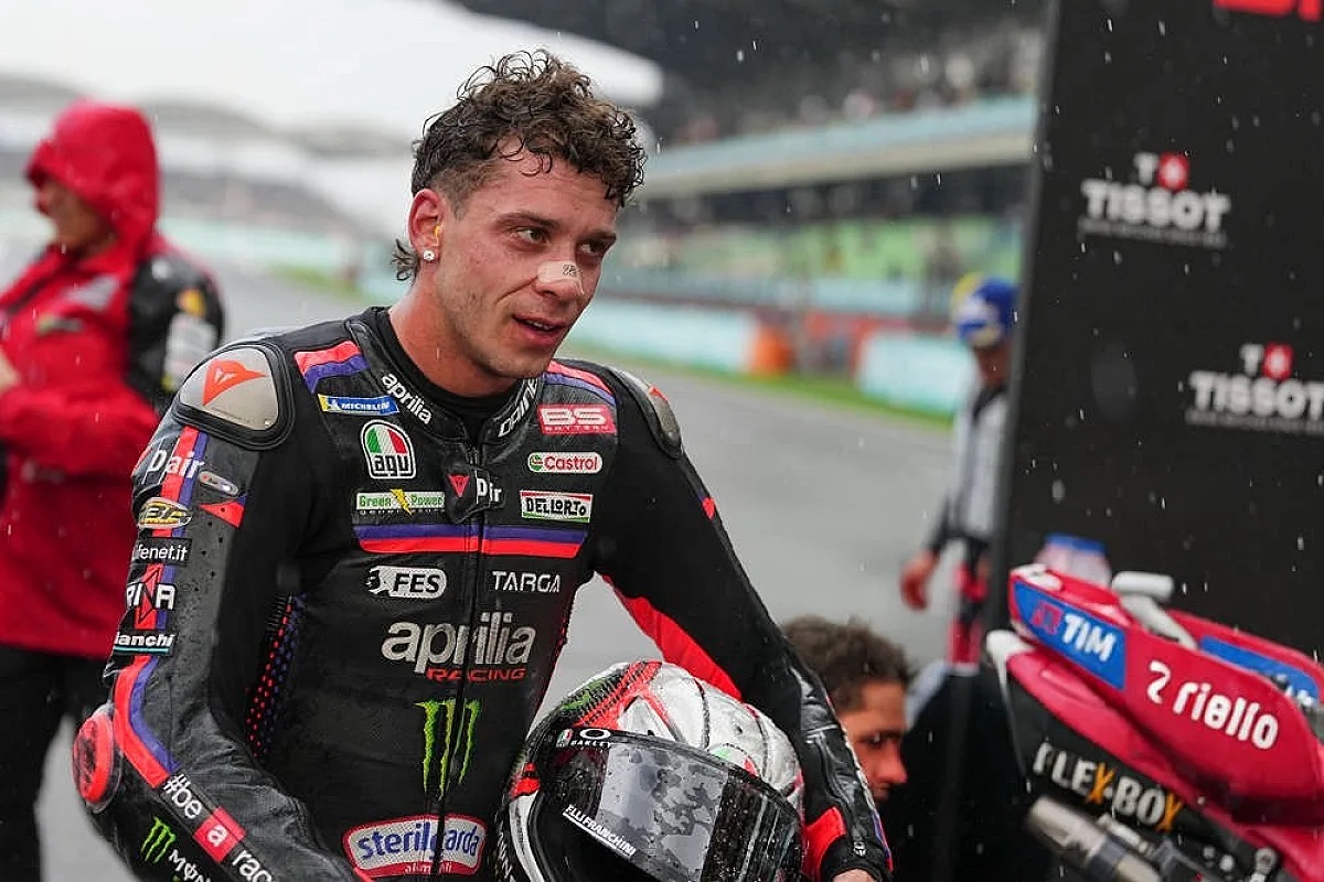 Rider in black Aprilia Racing leathers with a helmet under his arm, standing in the rain at the Sepang MotoGP paddock by a red bike