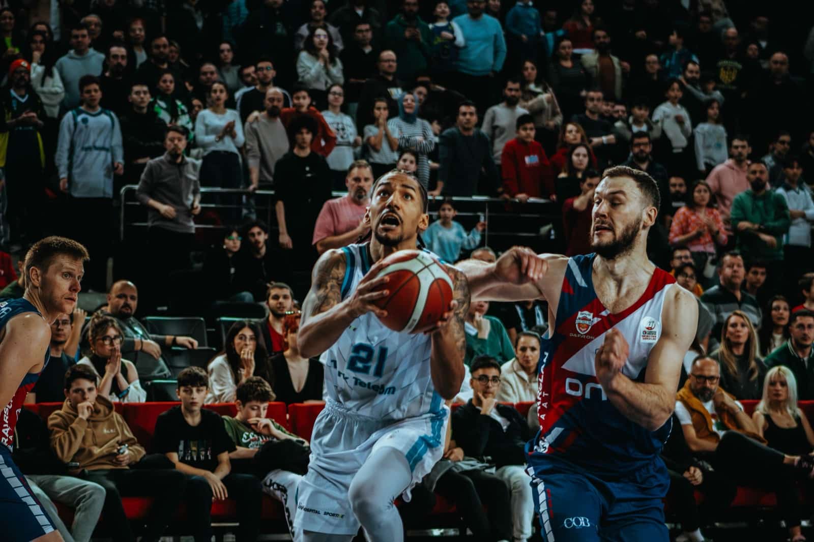 A basketball player drives toward the basket against a defender in front of a packed arena crowd