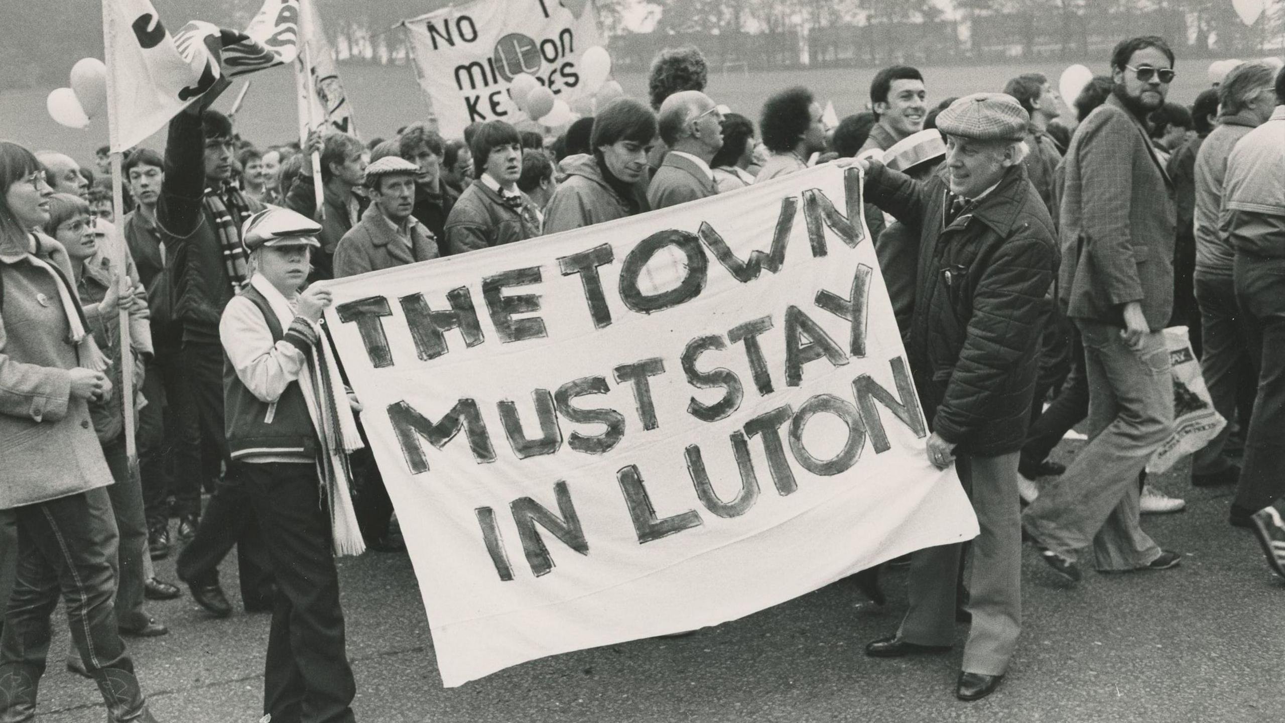 Luton Town fans are protesting. Two are holding a black and white sign which reads, 'the Town must stay in Luton.'