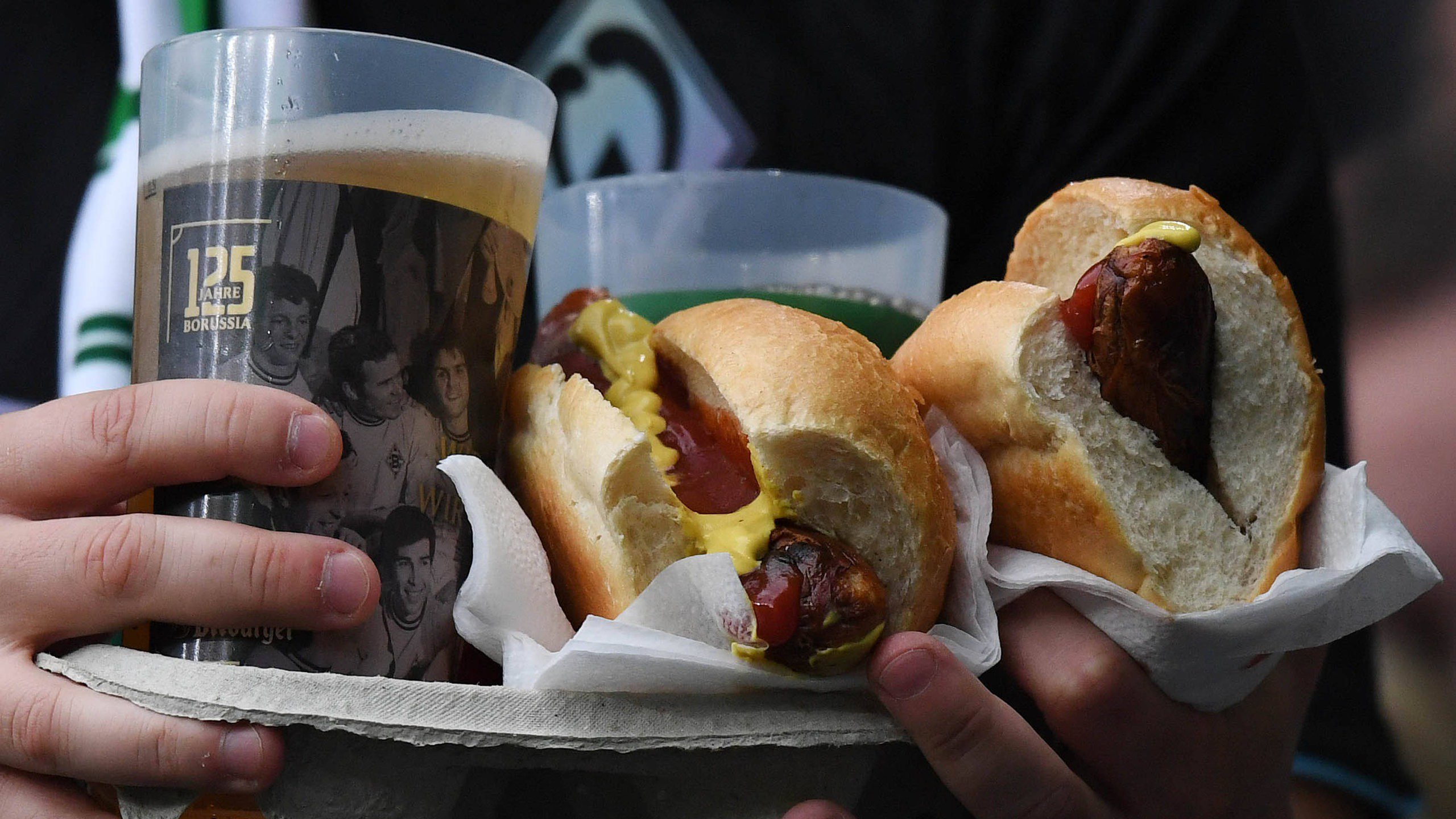 Nahaufnahme eines Werder-Fans, der im Stadion einen Teller mit zwei Bierbechern und zwei Bratwürsten im Brötchen in der Hand hält.