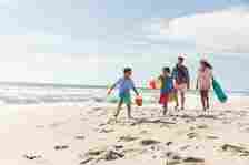Padres multirraciales caminando detrás de los niños corriendo en la arena de la playa durante el día soleado