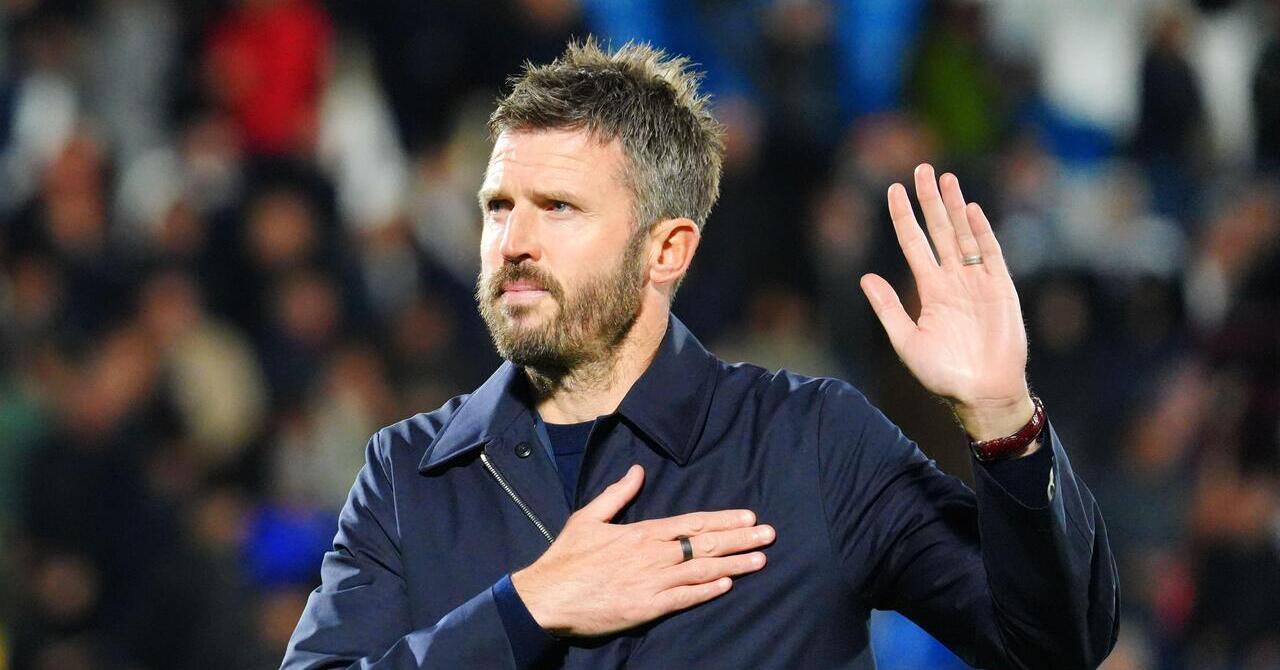 Football manager Michael Carrick looking upwards to his right in a stadium in his role as head coach of Manchester United Football Club. Carrick is wearing a black jacket over a dark blue top and holding his right hand to the left of his chest while raising his left hand.