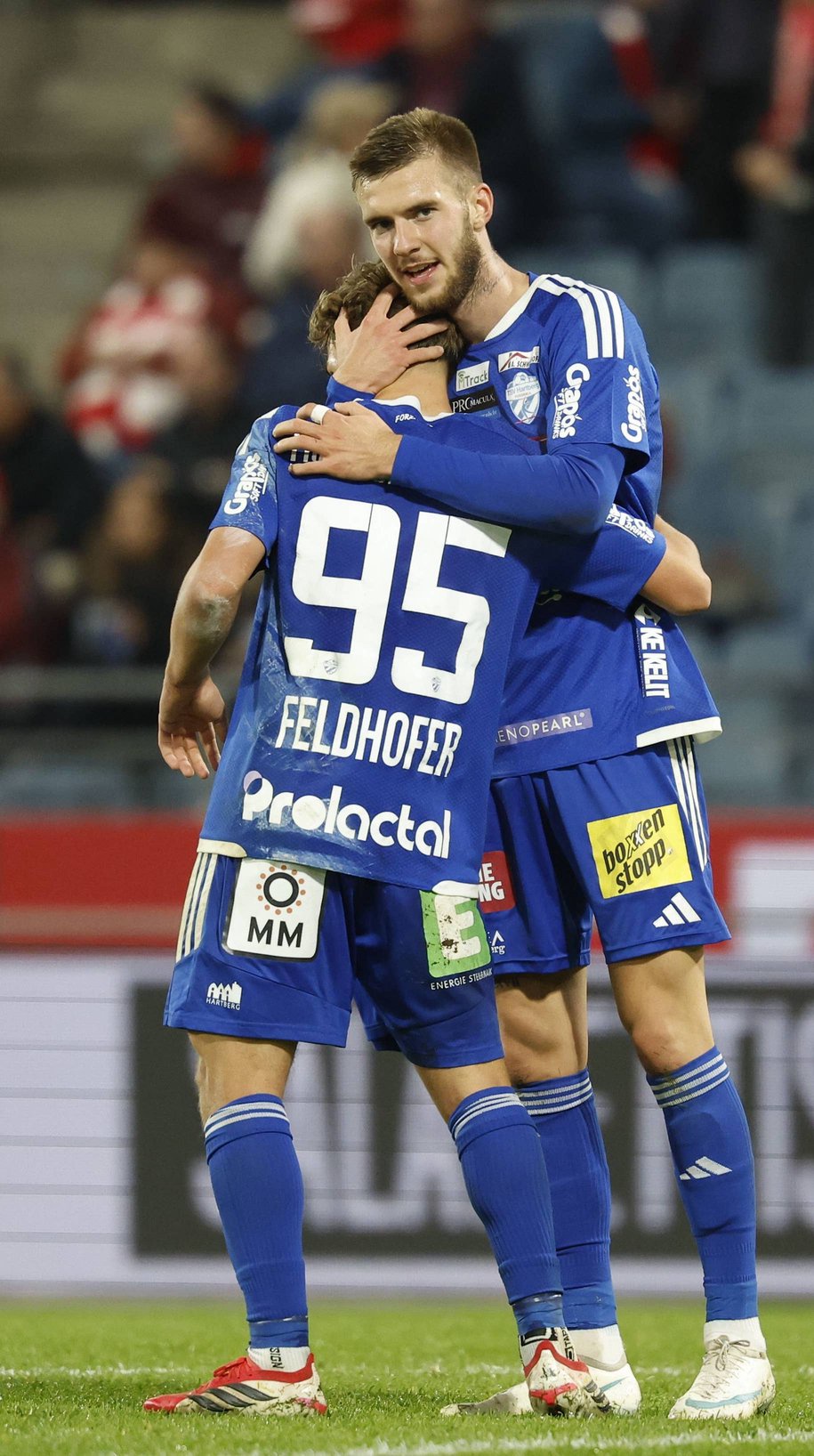 Hartberg-Torschütze Nikolaus Feldhofer im blauen Trikot mit der Rückennummer 95 umarmt jubelnd einen Teamkollegen Oliver Lukic auf dem Spielfeld.