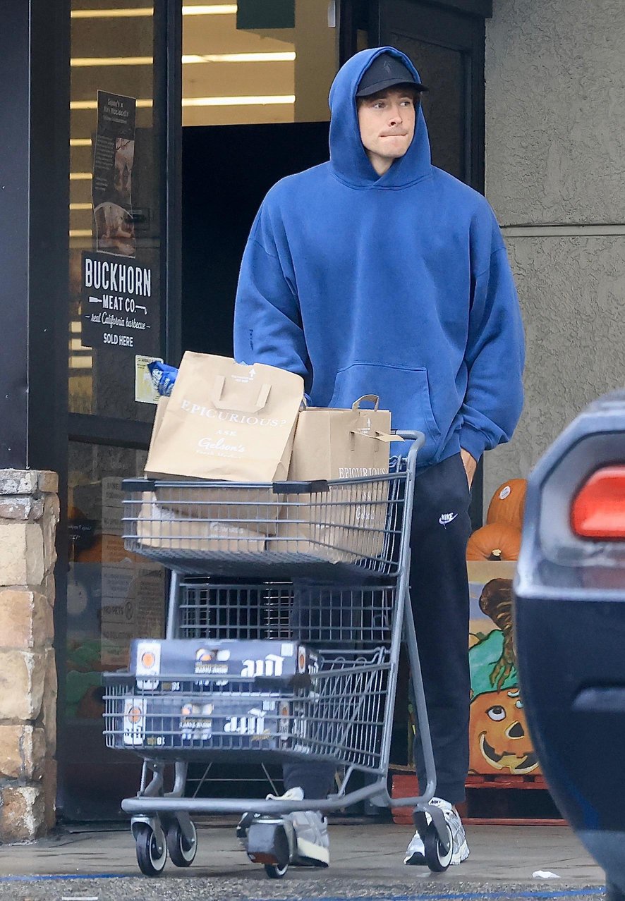 Justin Herbert wearing a blue hoodie and black pants stands with a shopping cart filled with groceries outside a store.