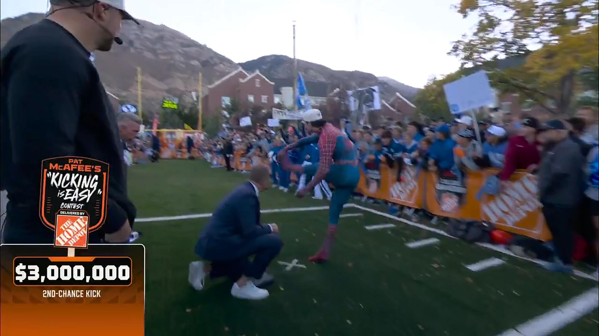 A person dressed in a Spider-Man suit prepares to kick a field goal on a football field, while a crowd watches and a man kneels nearby to hold the ball.