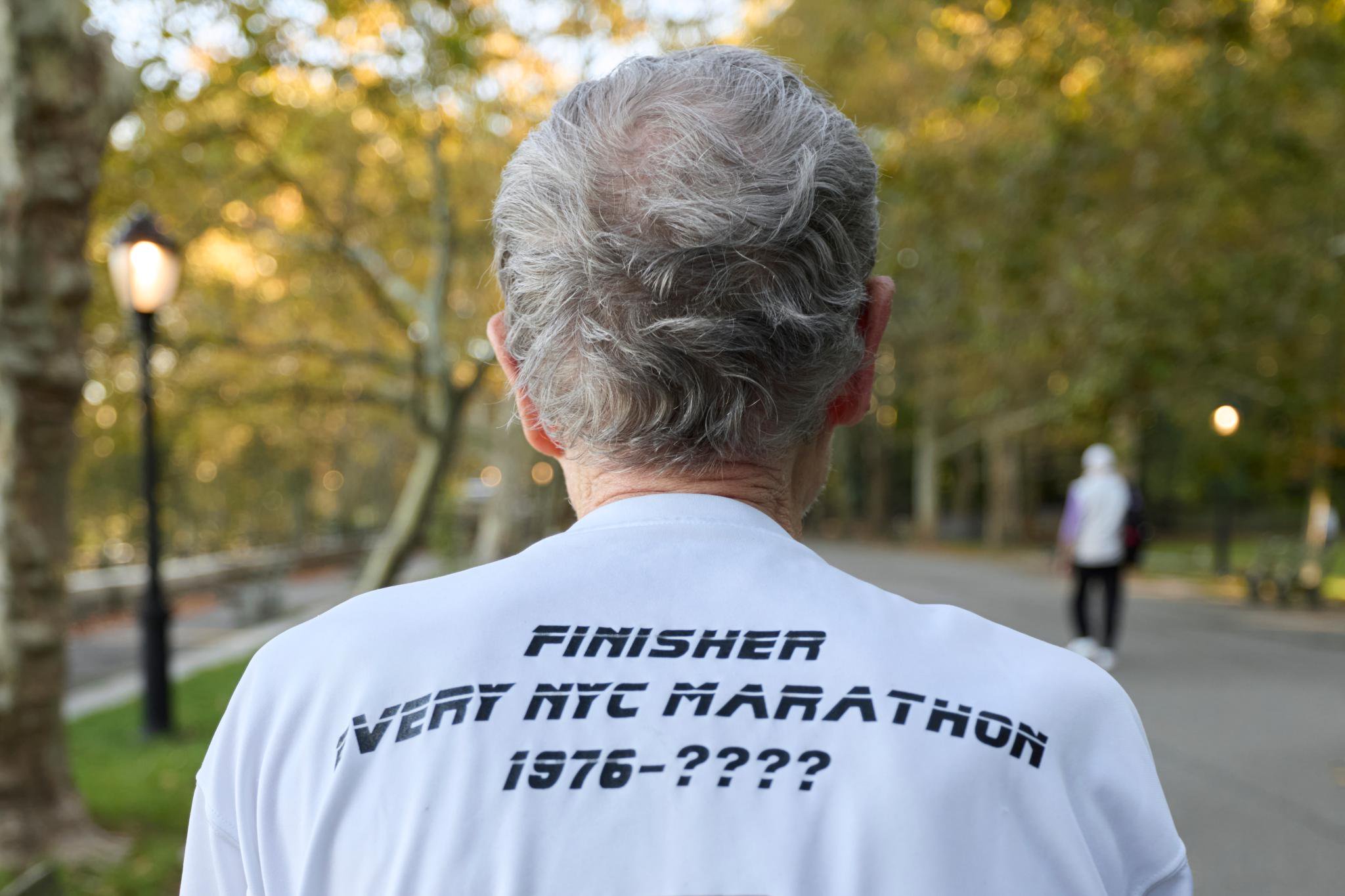Dave Obelkevich, the only person to complete every NYC Marathon since 1976, wears a shirt that reads "Finisher Every NYC Marathon 1976-????" while walking in Riverside Park.