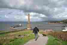 A man and a dog walk down a path with green grass on either side toward a chimney and the sea in front of them.
