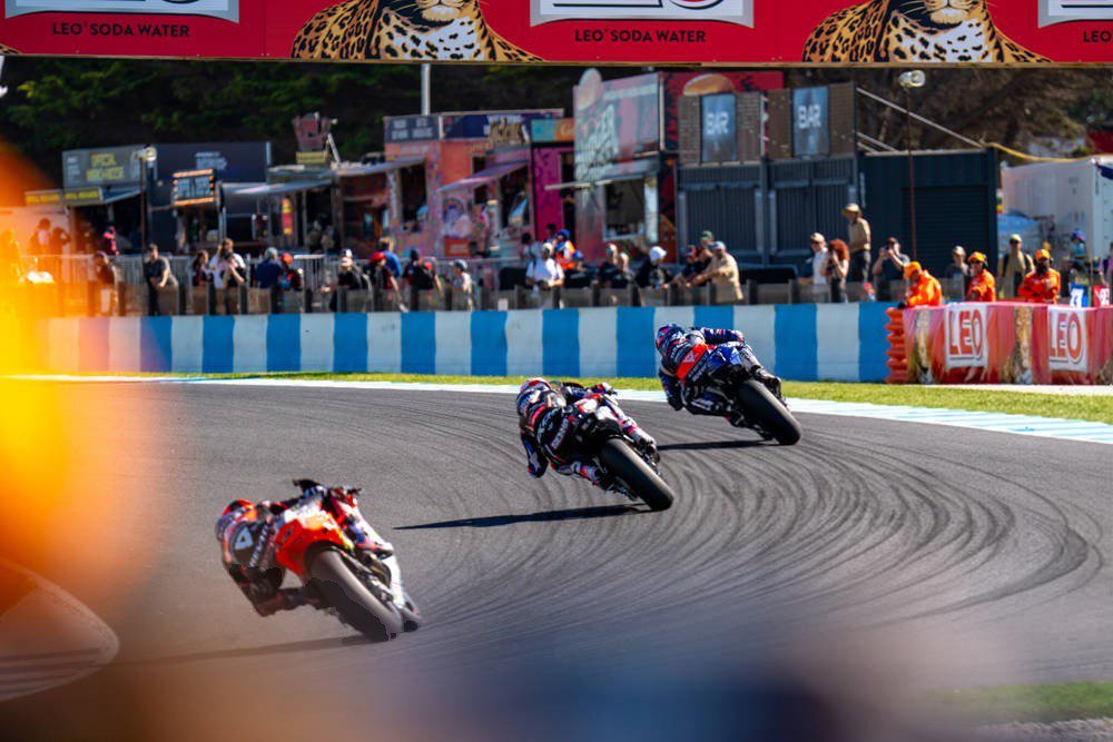 Two MotoGP riders lead a pack through a corner, one in red-and-white Ducati livery, the other on a dark blue Yamaha, at a MotoGP race.