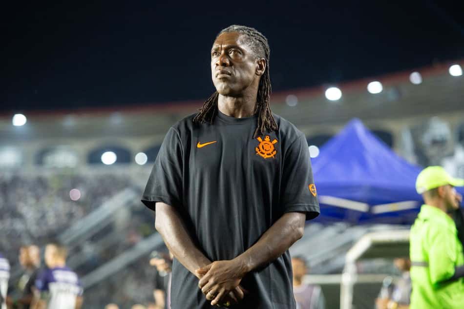 Homem com dreadlocks, camiseta preta com emblema laranja, braços cruzados em estádio durante evento.