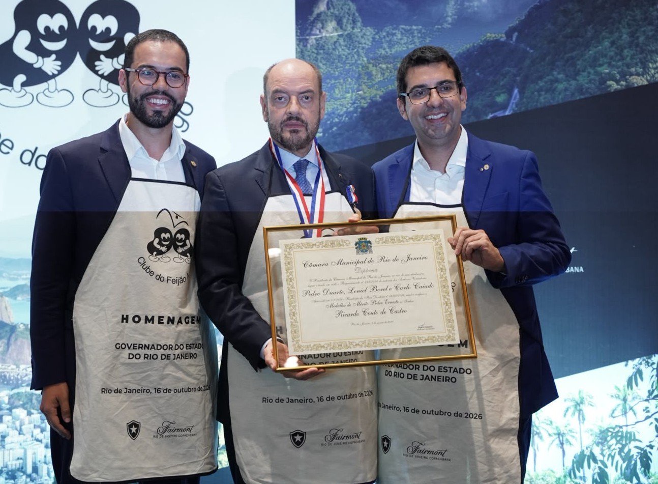Três homens sorridentes, de terno e aventais claros do Clube do Feijão, posam com um diploma emoldurado. O homem do centro usa uma medalha no pescoço. Ao fundo, uma projeção da cidade do Rio de Janeiro