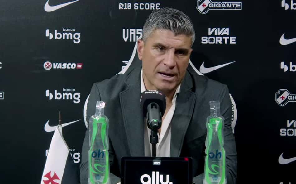 Treinador em coletiva de imprensa Vasco x Corinthians, diante de um microfone, com garrafas de água e backdrop com logos