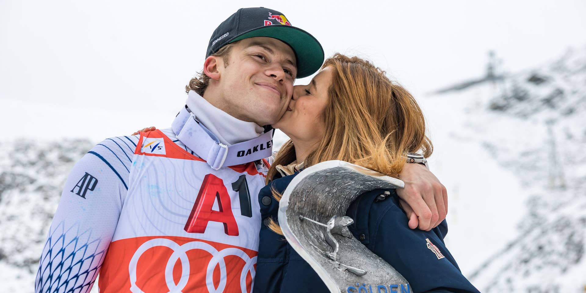 Ein Skifahrer wird bei der Siegerehrung von einer Frau auf die Wange geküsst, mit Pokal in der Hand.