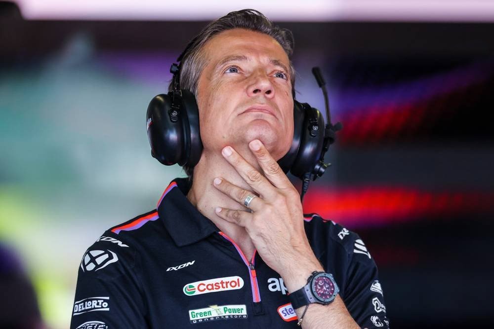 A man in a dark Aprilia team polo with a headset, hand on his chin, looking up in the MotoGP paddock with Castrol and Green Power sponsor logos visible
