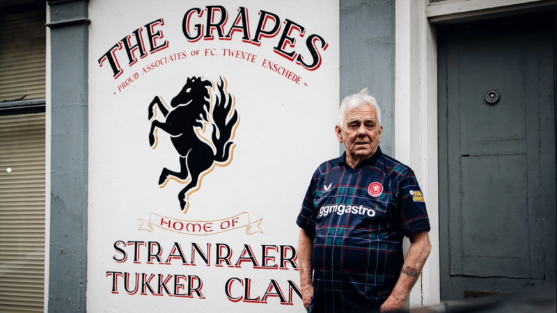 Billy Hodge standing outside The Grapes pub in Stranraer. It has the name of the bar in black text on a white background behind him and a large icon of a horse. He is wearing the shirt and looking slightly off camera.