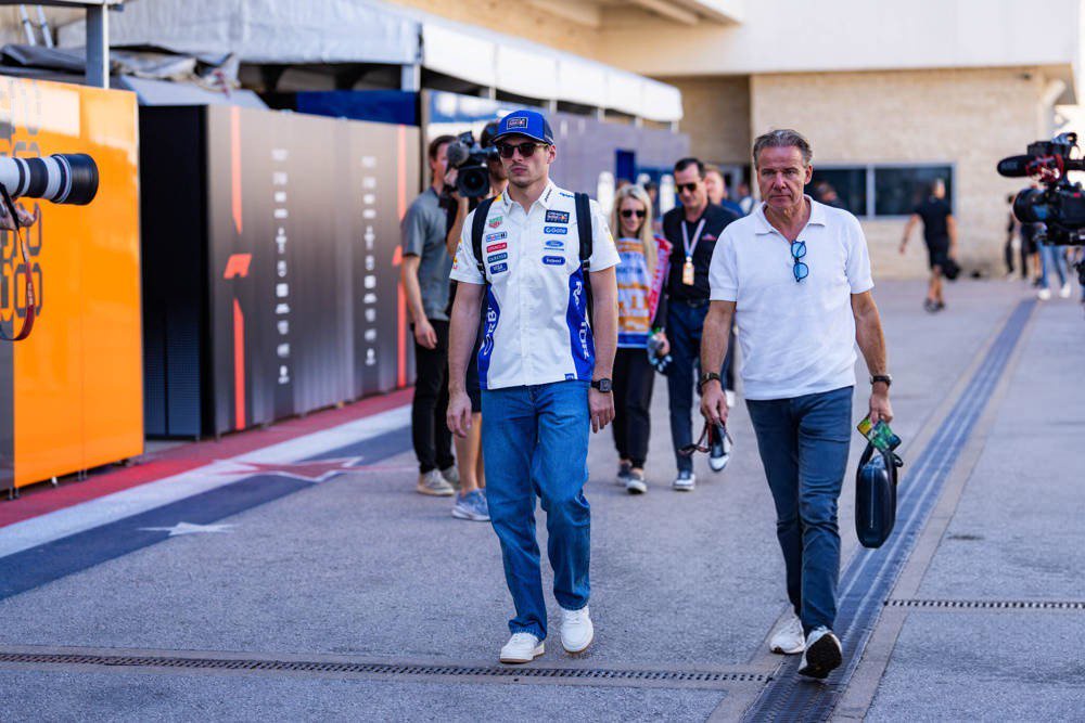 A Formula 1 driver in a white and blue team jacket and cap walks a paddock with a second man in a white shirt, past orange and dark garage panels and TV came…