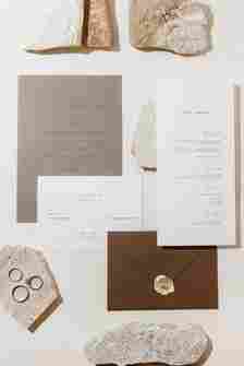 Minimal Brown and White Wedding Invitations Next to Rings and Rock Decor