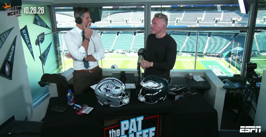 Two men broadcasting from a stadium press box, with a Dallas Cowboys helmet and a Philadelphia Eagles helmet on the desk in front of them.