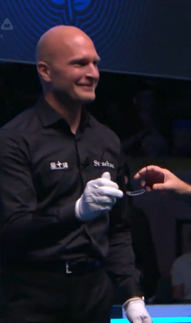 A bald referee wearing a black shirt and white gloves smiles while holding a white cue ball.