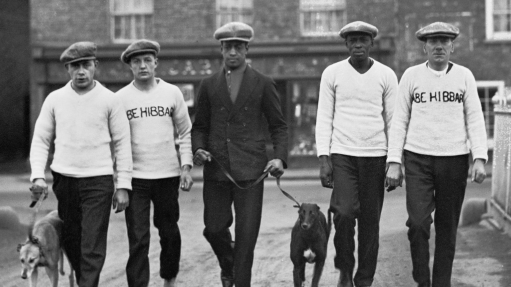 Johnson (centre), pictured with his father and manager Bill (second from right) in Manchester, December 1926. All five men wear flat caps and jumpers and walk with two greyhounds.