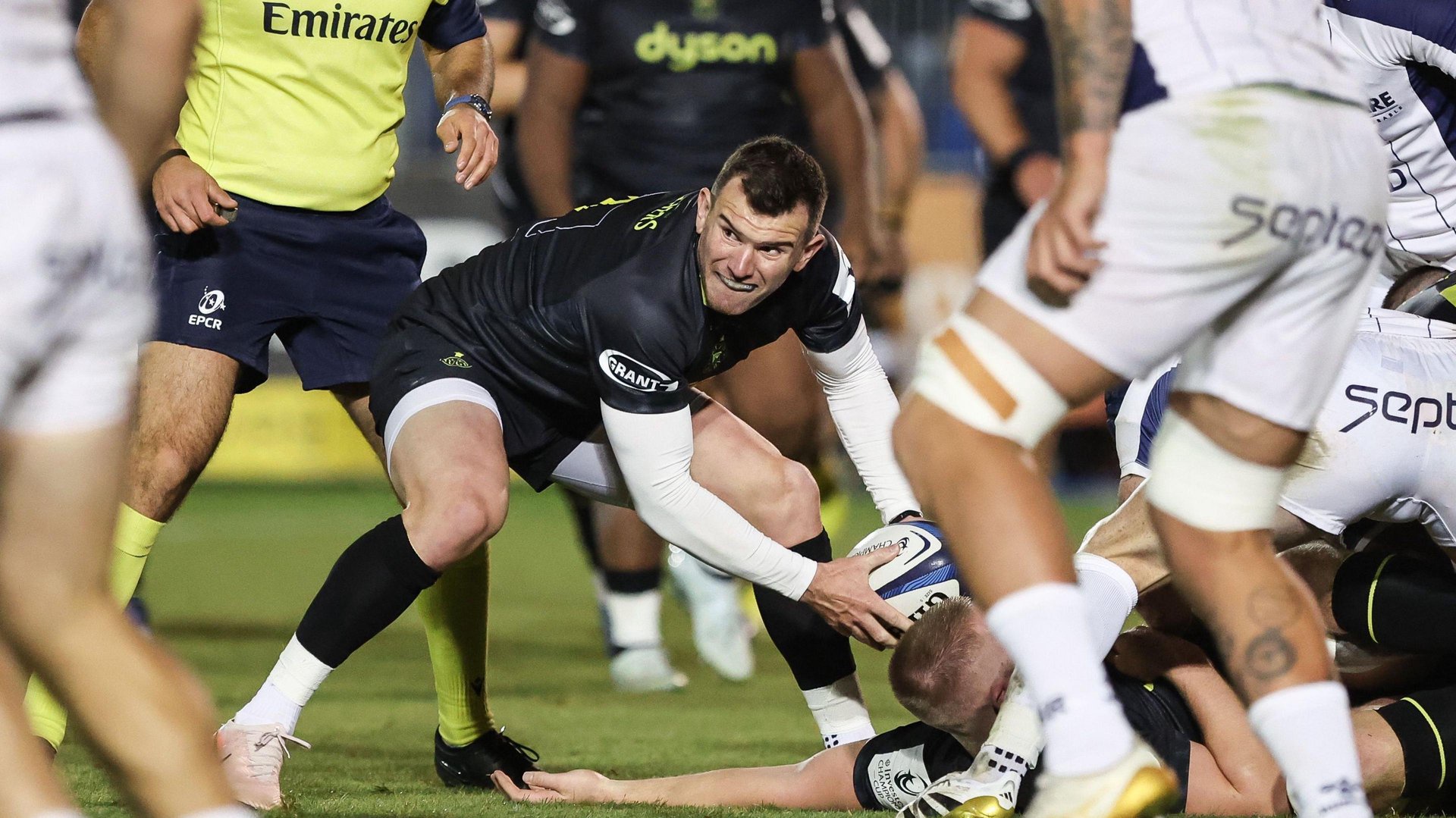 Ben Spencer crouches forward and holds a rugby ball in two hands as he picks it up off the floor during Bath v Montpellier. He is looking to his right and wearing black Bath kit