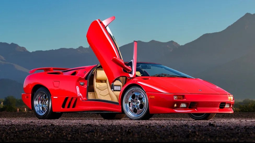 A red Lamborghini Diablo parked on pavement with the passenger door open in front of a distant mountain range