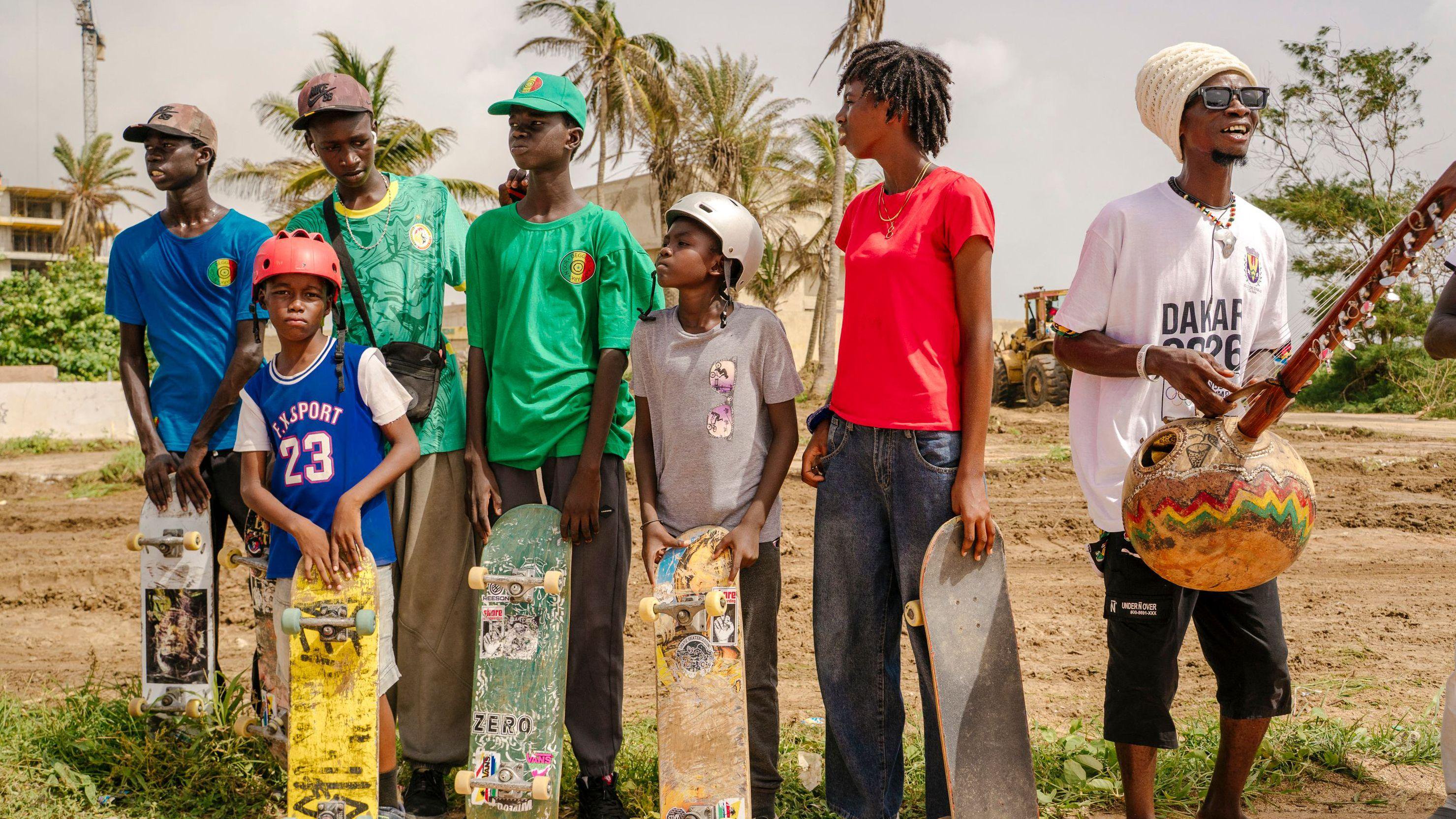 Six young skateboarders stand behind their boards, which largely rest upright in front of their legs, on a patch of land which is largely dirt with patches of scrubby green grass and palm trees in the background. The six wear a mix of colourful shirts, including a couple of green Senegal football shirts and a blue basketball top. Three wear baseball caps while the two smallest and youngest have helmets on. To the right of the five skateboards stands a man in a white Dakar 2026 T-shirt, sunglasses and a large knitted white hat who holds a traditional stringed instrument with a long neck and round, ball-like body
