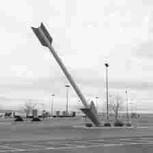 A giant arrow sculpture is embedded diagonally into the concrete in a mostly empty parking lot. The sky is cloudy, and there are a few barren trees and streetlights in the background.