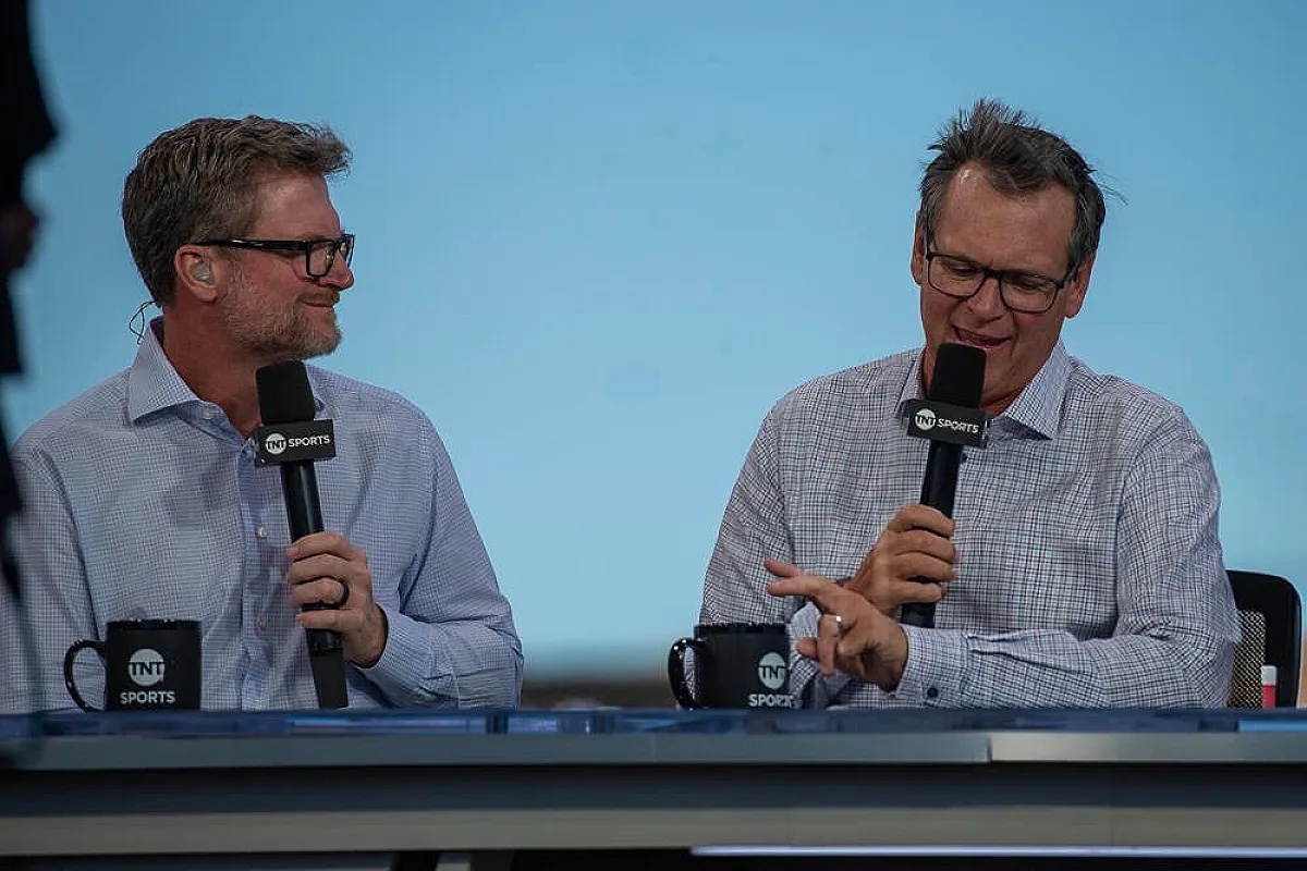 Two men in glasses and checked shirts at a TNT Sports broadcast desk, one gesturing while the other smiles and holds a microphone.
