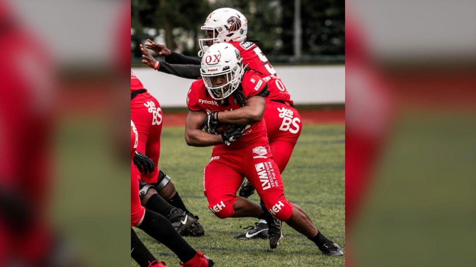 This is an action shot from an American football match. A man in a red kit and a white helmet holds a ball, surrounded by teammates.