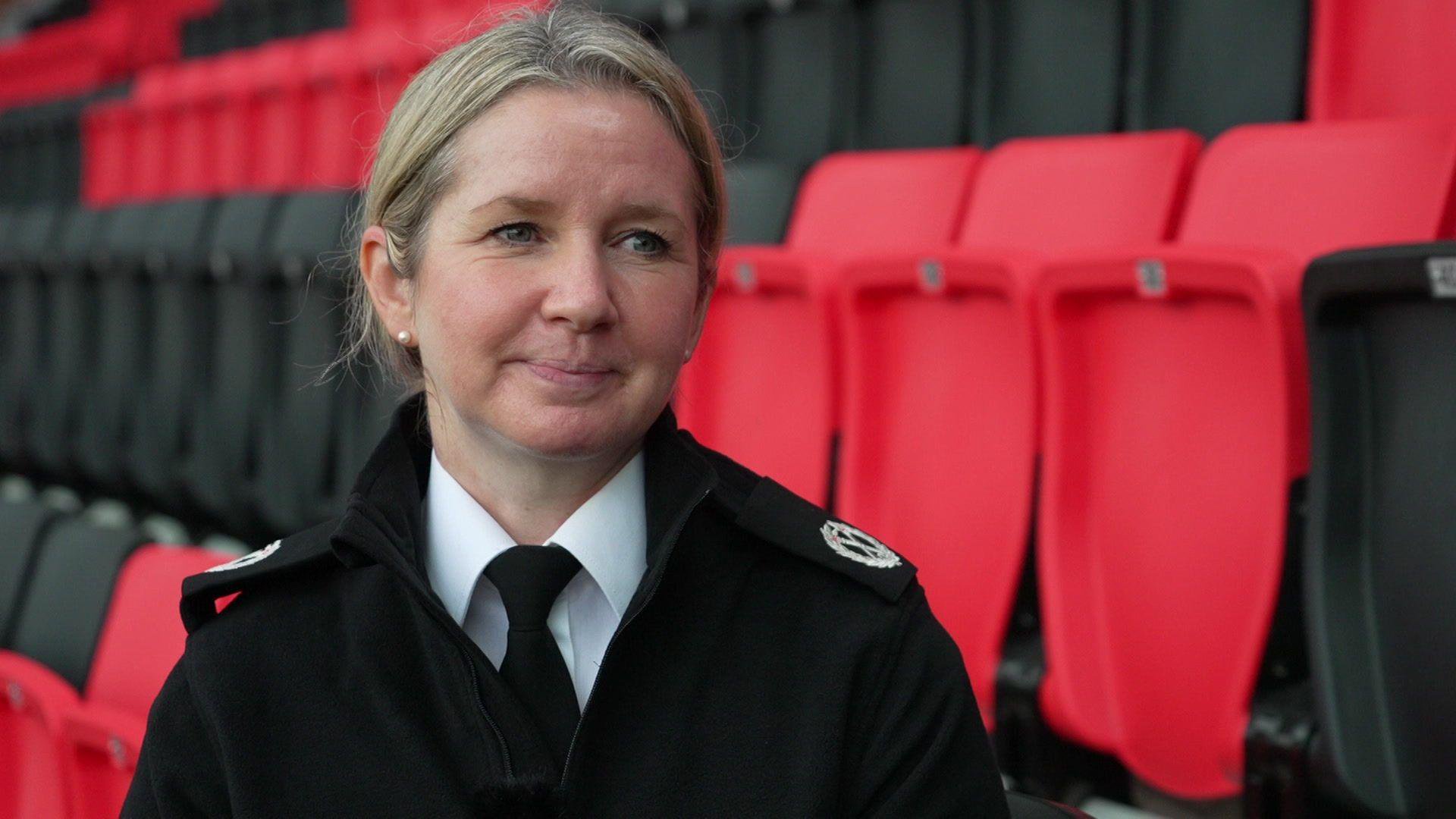 Assistant Chief Constable Lindsay Dudfield sits in the red and black stands of AFC Bournemouth for an interview. She has tied back blonde hair and is wearing  a black police uniform with a white shirt and black tie.