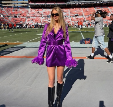 A blonde woman wearing a purple satin dress with feather cuffs and knee-high black boots stands on the field of a large football stadium.
