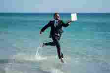 A businessman in a full suit and tie frolics across the beach with his laptop like a LinkedIn lunatic