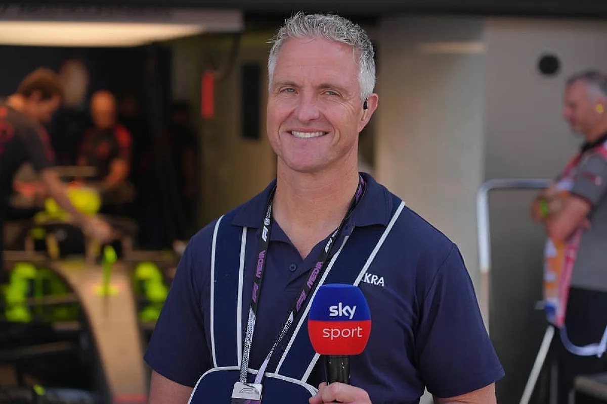 A smiling man with short grey hair, wearing a navy polo and headset, holds a Sky Sport microphone in a paddock area with blurred crew members behind him.