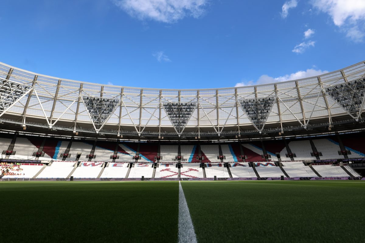 Empty London Stadium interior with green pitch, white sideline, and maroon/blue seating under a blue sky.