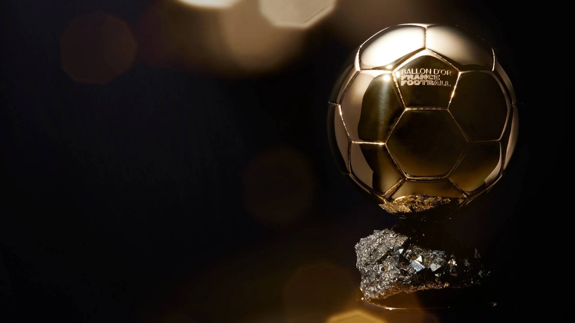 Troféu Bola de Ouro, dourado e brilhante, em formato de bola de futebol com gomos hexagonais, sobre uma base de pedras escuras e irregulares. O fundo é preto, com pontos de luz dourada desfocados