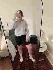 A person wearing a gray sweatshirt and black shorts takes a mirror selfie in a bedroom with a rug, bed, and side table.