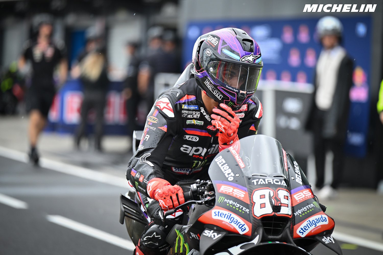 A rider in a purple helmet and Aprilia leathers on an Aprilia MotoGP bike with number 89 in the pit lane, gesturing as he rides past the garages.